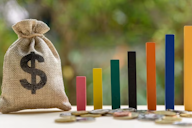 A burlap sack with a dollar sign next to colourful vertical blocks and scattered coins, symbolising budgeting and financial growth.